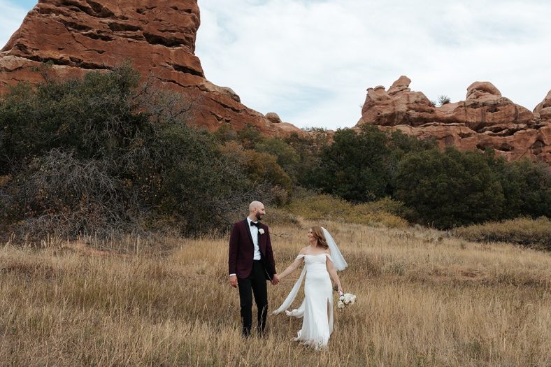 A bride and groom hold hands and walk through tall golden grass in a scenic Colorado landscape, framed by dramatic red rock formations. The bride wears an elegant off-the-shoulder white gown with a veil and holds a bouquet of white flowers, while the groom is dressed in a burgundy tuxedo jacket and black trousers. The couple looks at each other, creating a romantic and adventurous wedding portrait set against the natural beauty of the mountains.
