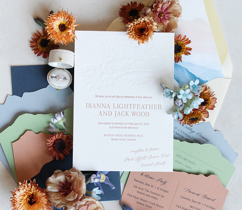 A mountain-inspired wedding invitation suite featuring a white letterpress main invite with names in terracotta ink, surrounded by die-cut detail cards in green, terracotta, and slate blue. The suite is styled for a romantic Beaver Creek, Colorado celebration.