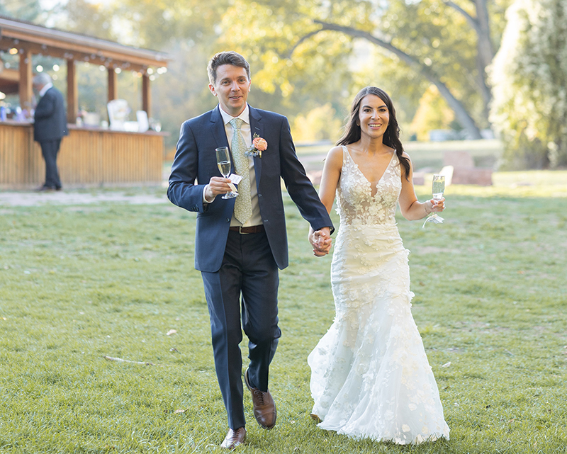 Bride and groom walk hand in hand with champagne glasses during their outdoor wedding reception at Planet Bluegrass in Lyons, Colorado.