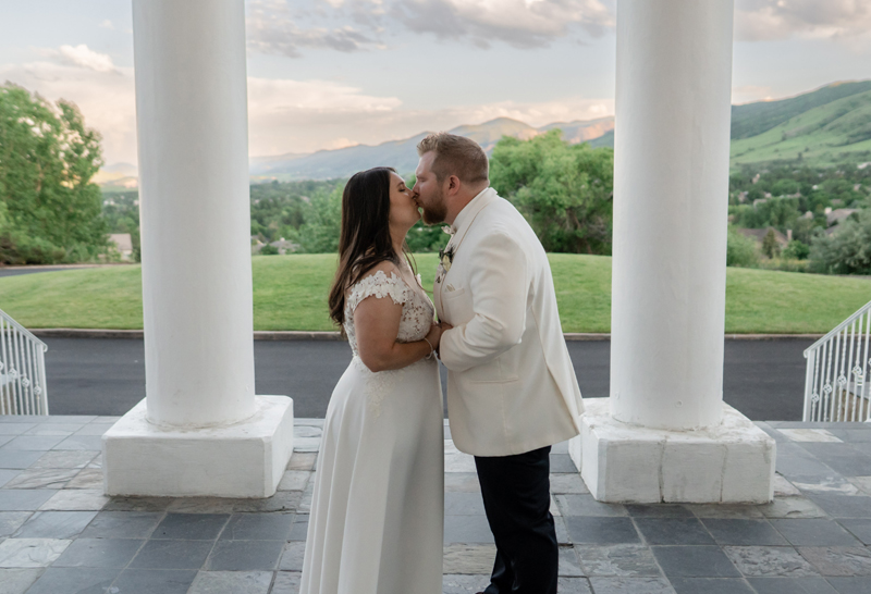Bride and groom share a kiss between the white columns of The Manor House in Littleton, Colorado, with foothill views behind them at sunset.